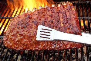 Large rack of BBQ ribs being held by a pair of metal silver tongs on a family grill for a BBQ in Springfield, IL.