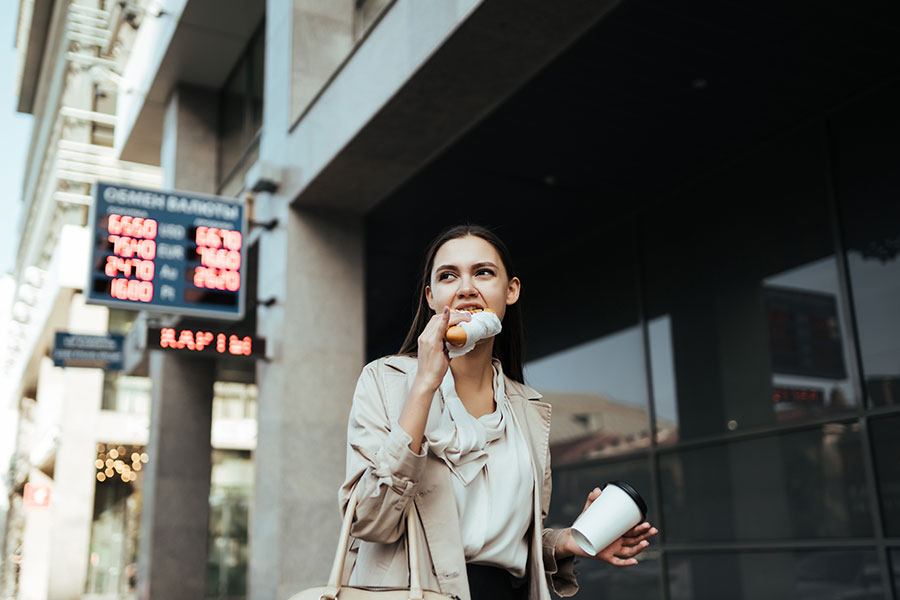 A woman in business clothing eating food on the go while holding a coffee in her hand and walking down a city street in Springfield, IL.