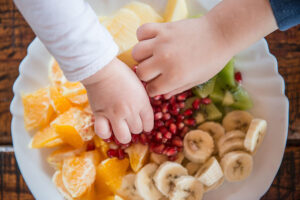 Two small hands reaching for healthy fruit like oranges, bananas, kiwi, and pomegranate seeds as daycare and after-school snacks in Springfield, IL.